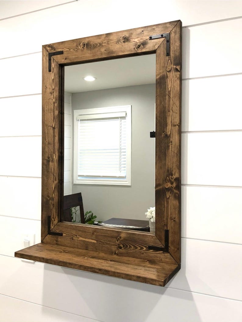 May include: A rectangular mirror with a dark brown wooden frame and a shelf. The frame has black metal corner brackets. The mirror is mounted on a white wall with horizontal panelling.