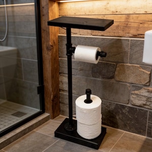 May include: A black metal toilet paper stand with a shelf. The stand holds a roll of toilet paper and has a base that holds two additional rolls. The stand is in a bathroom with a stone tile wall and a glass shower door.
