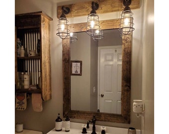 May include: A rectangular wall mirror with a dark stained wood frame, mounted above a white bathroom sink. Three industrial-style light fixtures with metal cages are attached to a matching wooden board above the mirror, illuminating the space.