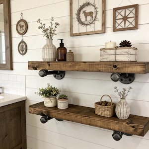 May include: Rustic bathroom shelves made of dark stained wood, supported by black pipe fittings. The shelves are decorated with vases, plants, and decorative items, creating a farmhouse style.