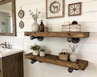 May include: Rustic bathroom shelves made of dark stained wood, supported by black pipe fittings. The shelves are decorated with vases, plants, and decorative items, creating a farmhouse style.
