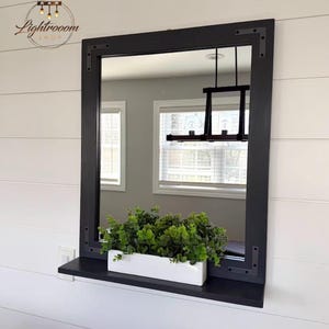 May include: A rectangular black framed mirror with a shelf, mounted on a white wall. A white rectangular planter filled with green plants sits on the shelf. The mirror reflects a room with windows and a black light fixture.