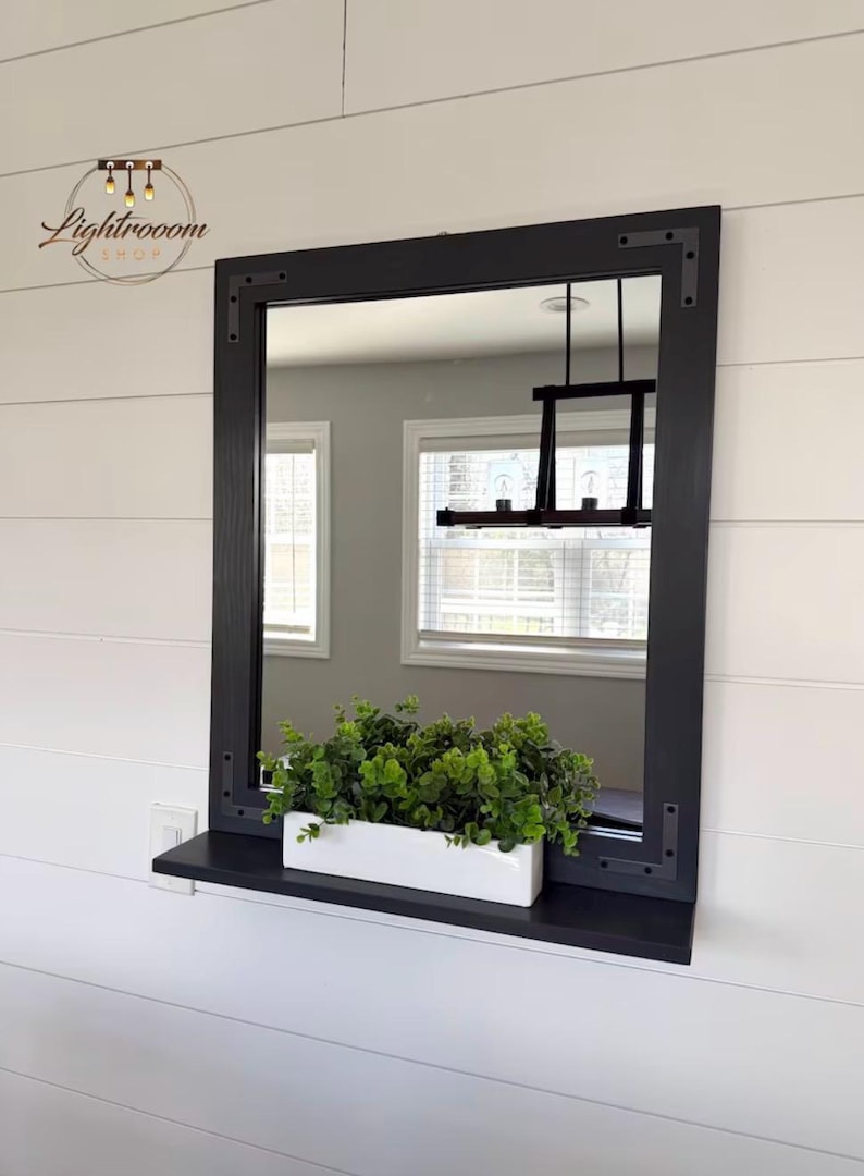 May include: A rectangular black framed mirror with a shelf, mounted on a white wall. A white rectangular planter filled with green plants sits on the shelf. The mirror reflects a room with windows and a black light fixture.
