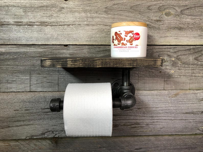 Puede incluir: Un estante de madera te&ntilde;ida oscura montado con un soporte para papel higi&eacute;nico de tuber&iacute;a industrial negra. Un rollo de papel higi&eacute;nico blanco est&aacute; en el soporte y una vela blanca con la etiqueta Gingerbread Cookies est&aacute; sobre el estante.