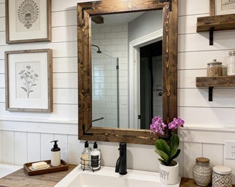 May include: A rectangular wall mirror with a dark stained wooden frame featuring black metal corner accents. The mirror is mounted above a white ceramic sink basin with a black faucet, positioned on a wooden countertop with various bathroom accessories.