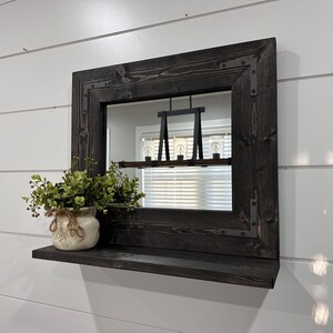 May include: A dark-stained wooden framed mirror with a shelf. The mirror has metal corner accents. A small jar with greenery sits on the shelf. The mirror reflects a light fixture and a window with blinds.