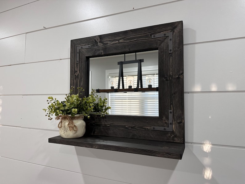 May include: A dark-stained wooden framed mirror with a shelf. The mirror has metal corner accents. A small jar with greenery sits on the shelf. The mirror reflects a light fixture and a window with blinds.