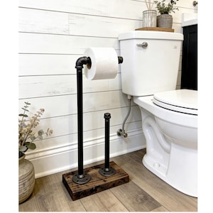 May include: A freestanding toilet paper holder constructed from black industrial metal piping and fittings, mounted onto a rectangular dark stained wooden base. The design features a tall vertical pipe for the roll and a shorter pipe for storage.