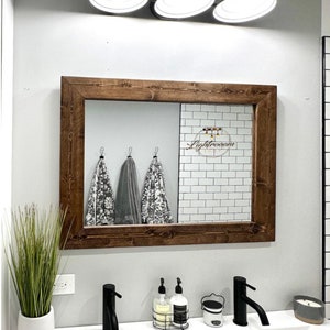 May include: A bathroom mirror with a dark brown wooden frame. The mirror reflects a white brick wall and three hanging towels. The word "Lightroom" is written on the mirror. Black faucets and a plant in a white pot are in the foreground.