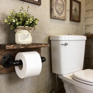 May include: A rustic wooden toilet paper holder with a shelf, featuring black pipe fittings and a roll of white toilet paper. A small white jar with the word "Ball" on it and greenery sits on the shelf. The holder is mounted on a textured wall.