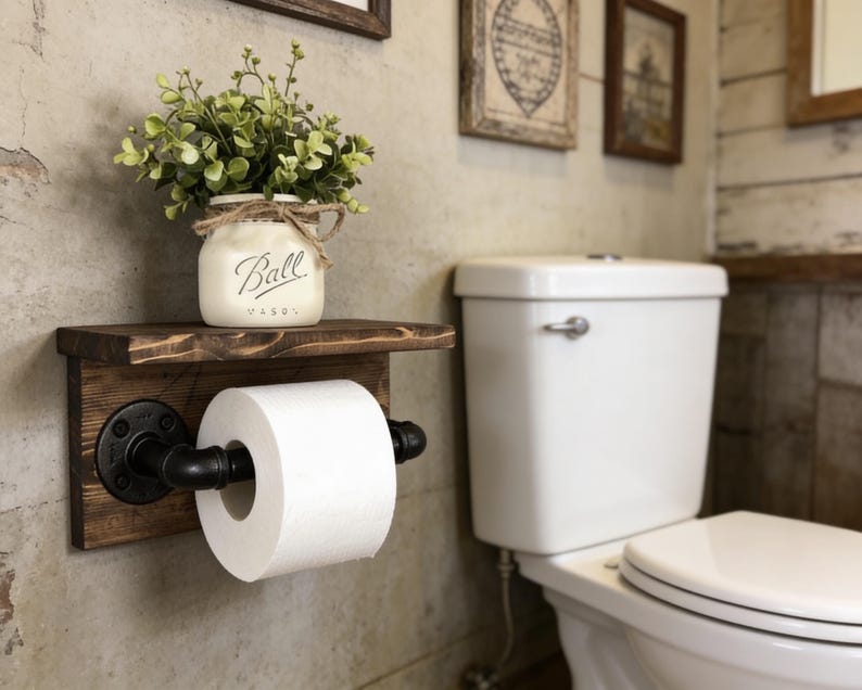 May include: A rustic wooden toilet paper holder with a shelf, featuring black pipe fittings and a roll of white toilet paper. A small white jar with the word "Ball" on it and greenery sits on the shelf. The holder is mounted on a textured wall.