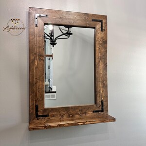 May include: A rectangular mirror with a dark brown wooden frame and a shelf. The frame has visible wood grain and black corner brackets. The mirror reflects a chandelier and a room's interior.