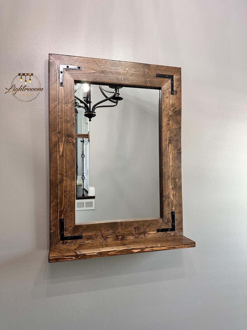 May include: A rectangular mirror with a dark brown wooden frame and a shelf. The frame has visible wood grain and black corner brackets. The mirror reflects a chandelier and a room's interior.