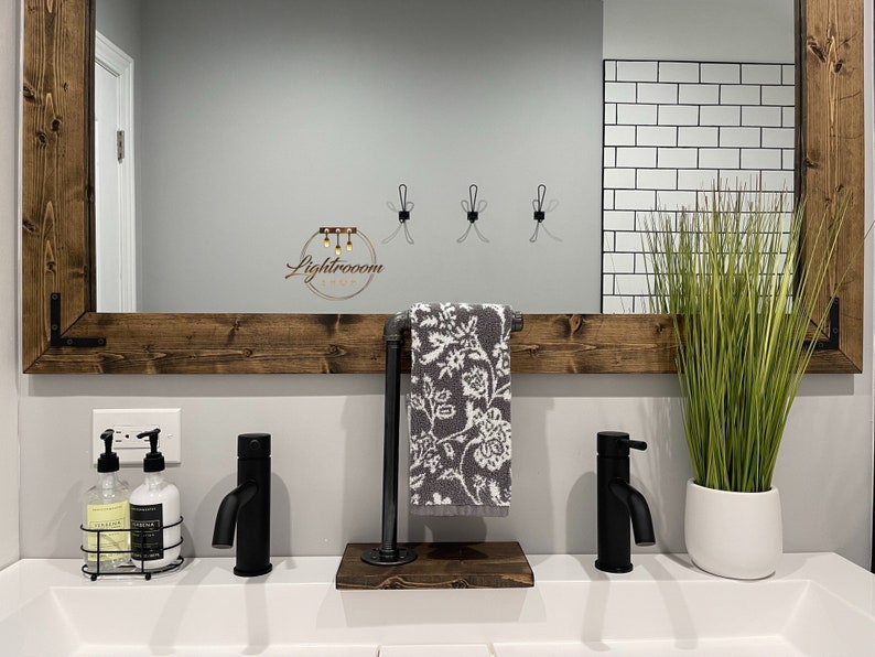 May include: A bathroom vanity setup featuring a dark wood framed mirror, two matte black faucets, and a gray and white floral hand towel hanging on a dark metal pipe stand with a wooden base. Includes two soap dispensers and a potted green plant.