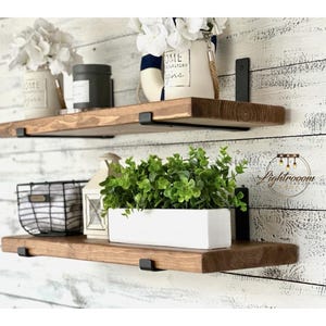 May include: Two horizontal wooden shelves with dark brown wood grain and black metal brackets. The top shelf holds a white vase with white flowers and a black container. The bottom shelf holds a black wire basket, a white lantern, and a white rectangular planter with green foliage.