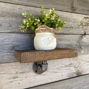 May include: A rustic wooden shelf with a dark stain, supported by a metal pipe fitting. A white Ball Mason jar with greenery and twine sits on the shelf, creating a farmhouse aesthetic. The shelf is mounted on a weathered wood wall.