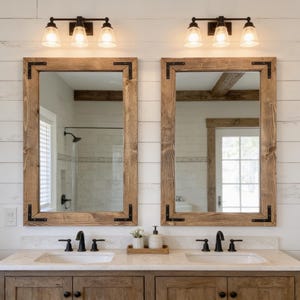 May include: Two rectangular mirrors with medium-toned wood frames and black metal corner brackets. Each mirror is mounted above a white sink with a black faucet, set into a wooden vanity cabinet against a white horizontal plank wall.