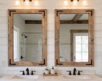 May include: Two rectangular mirrors with medium-toned wood frames and black metal corner brackets. Each mirror is mounted above a white sink with a black faucet, set into a wooden vanity cabinet against a white horizontal plank wall.