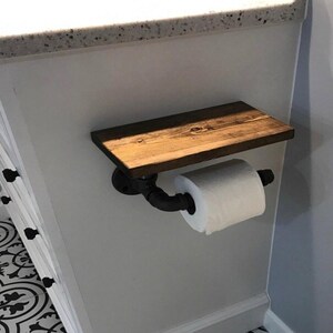 Puede incluir: Un portarrollos de papel higi&eacute;nico con un estante rectangular de madera te&ntilde;ida de marr&oacute;n oscuro montado sobre un soporte de tuber&iacute;a industrial negro. El soporte se extiende hacia afuera para sostener un rollo de papel higi&eacute;nico blanco.