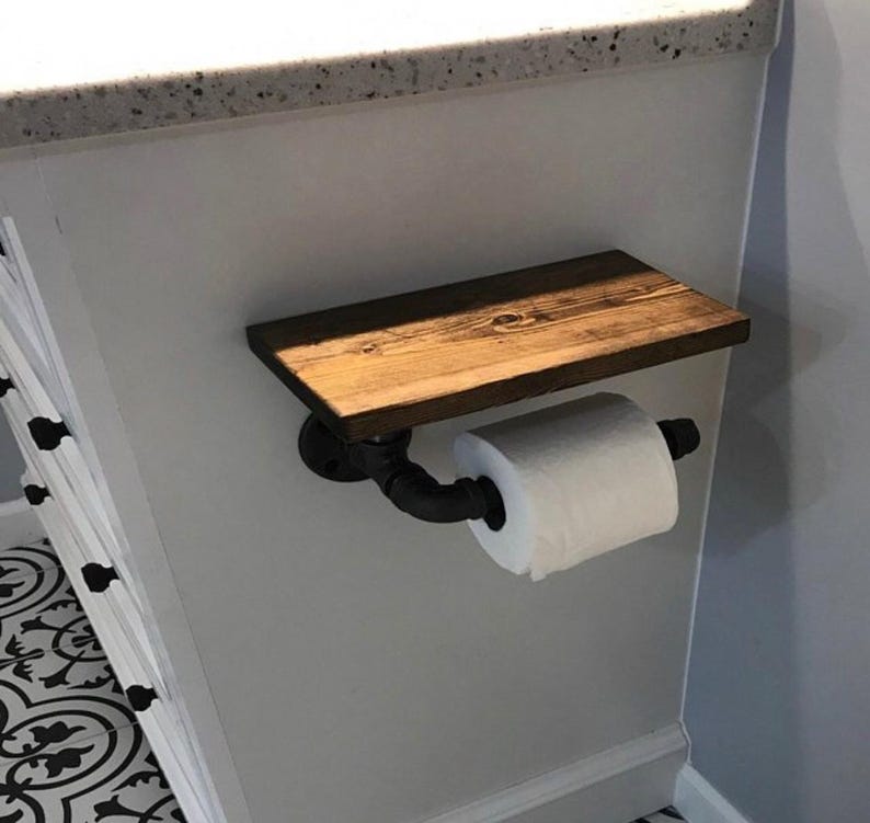 Puede incluir: Un portarrollos de papel higi&eacute;nico con un estante rectangular de madera te&ntilde;ida de marr&oacute;n oscuro montado sobre un soporte de tuber&iacute;a industrial negro. El soporte se extiende hacia afuera para sostener un rollo de papel higi&eacute;nico blanco.