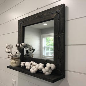 May include: A square, dark-stained wooden framed mirror with a shelf, mounted on a white wall. The shelf holds cotton stems in a mason jar and several cotton bolls. The mirror reflects a window and interior space.