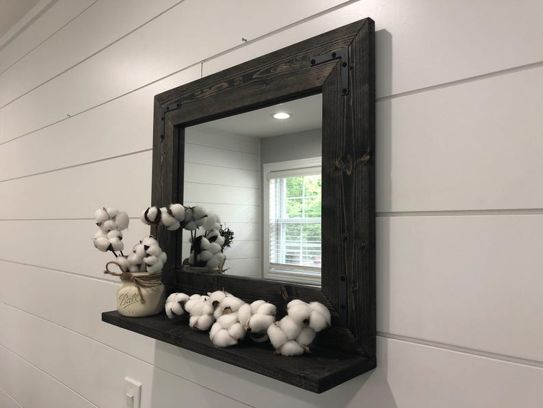 May include: A square, dark-stained wooden framed mirror with a shelf, mounted on a white wall. The shelf holds cotton stems in a mason jar and several cotton bolls. The mirror reflects a window and interior space.