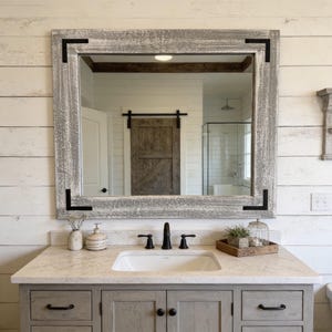 May include: A rectangular wall mirror with a weathered gray wood frame featuring black metal corner brackets. The mirror is mounted above a bathroom vanity with a white countertop, a white sink, and a gray cabinet with black hardware.