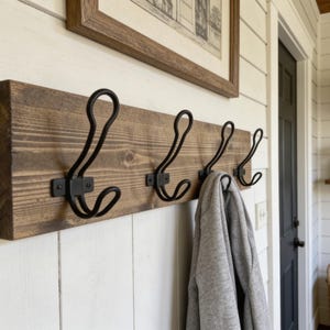 May include: A rectangular wooden wall-mounted coat rack featuring four black metal hooks with a double-prong design. The wood has a medium-brown stained finish with visible grain patterns. A grey hooded sweatshirt hangs from one of the hooks.