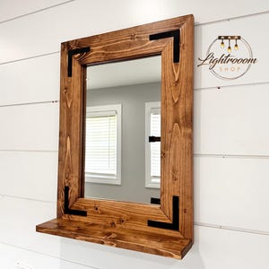 May include: A rectangular wooden mirror with a shelf, stained in a warm brown hue. The frame features black corner accents. The mirror is mounted on a white wall. The Lightrooom Shop logo is in the upper right corner.