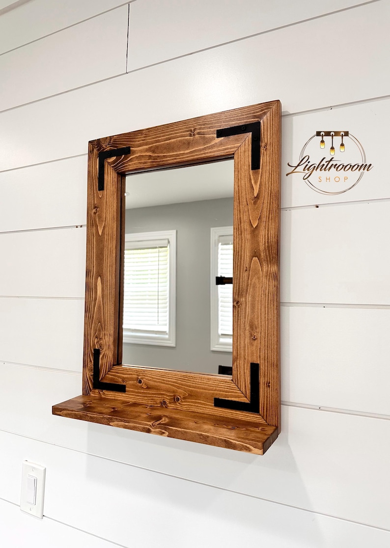May include: A rectangular wooden mirror with a shelf, stained in a warm brown hue. The frame features black corner accents. The mirror is mounted on a white wall. The Lightrooom Shop logo is in the upper right corner.