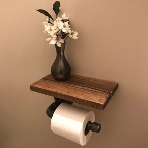 Puede incluir: Un soporte de papel higi&eacute;nico montado en la pared con un estante rectangular de madera marr&oacute;n oscuro sostenido por tuber&iacute;as industriales de metal negro. Un rollo de papel higi&eacute;nico blanco est&aacute; montado en el tubo y un jarr&oacute;n oscuro con flores blancas descansa encima.
