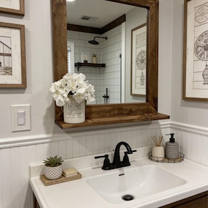 May include: A bathroom vanity with a white sink, black faucet, and a large wooden-framed mirror. The mirror reflects a shower and decorative items. A white ceramic pot with the word "HOME" and white flowers sits on a shelf below the mirror.