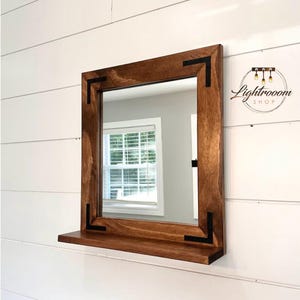 May include: Square wooden mirror with a dark brown frame and a small shelf. The frame has black corner accents. The mirror is mounted on a white shiplap wall. The Lightrooom Shop logo is in the upper right corner.