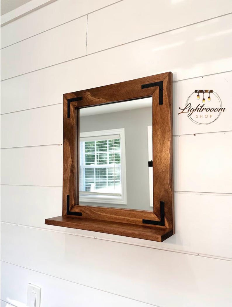 May include: Square wooden mirror with a dark brown frame and a small shelf. The frame has black corner accents. The mirror is mounted on a white shiplap wall. The Lightrooom Shop logo is in the upper right corner.