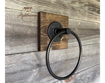 May include: A wall-mounted towel ring featuring a circular black metal ring attached to a square, dark-stained wooden base. The hardware includes a black circular flange secured with four screws. The logo Lightrooom Shop is visible in the upper left.