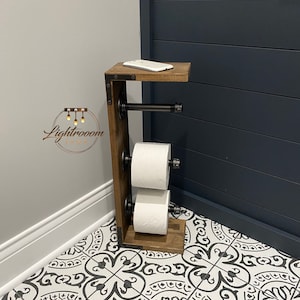 May include: A rustic wooden toilet paper holder with a shelf, holding two rolls of white toilet paper. The holder has black metal pipe accents and a small shelf with a white phone. The design is industrial and functional.