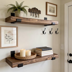 May include: Two dark stained wooden floating shelves with black metal brackets are mounted on a light beige wall. The top shelf holds a potted fern and a key rack. The bottom shelf displays candles, a woven tray, and books. Framed art pieces are also present.