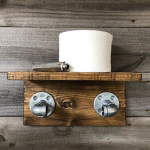 May include: A rustic, wooden toilet paper holder with a shelf. The shelf holds a roll of white toilet paper and a silver object. The holder features two metal pipe fittings and is mounted on a wooden plank.