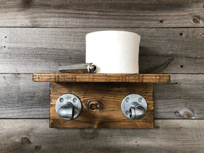 May include: A rustic, wooden toilet paper holder with a shelf. The shelf holds a roll of white toilet paper and a silver object. The holder features two metal pipe fittings and is mounted on a wooden plank.