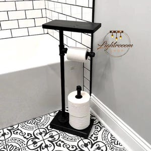 May include: Black metal toilet paper stand with a small shelf. The stand holds one roll of toilet paper and has a base to hold extra rolls. The stand is in a bathroom with black and white patterned tile.