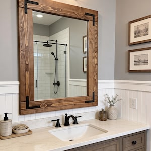 May include: A rectangular wooden framed mirror with black metal corner brackets hangs above a bathroom vanity. The mirror reflects a shower with a black shower head. The vanity has a white countertop and a dark brown cabinet.