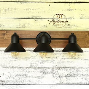 May include: A three-light vanity fixture with black metal shades and a dark wood base. The fixture has a rustic, industrial design, with exposed light bulbs. The fixture is mounted on a white and light brown wooden wall.
