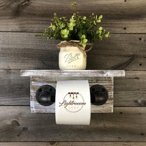 May include: Rustic wooden bathroom shelf with a distressed white finish, featuring a toilet paper holder with black pipe fittings. A Ball Mason jar with greenery sits on the shelf. The toilet paper roll has the text "Lightroom Shop".