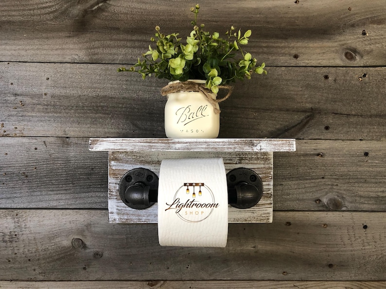 May include: Rustic wooden bathroom shelf with a distressed white finish, featuring a toilet paper holder with black pipe fittings. A Ball Mason jar with greenery sits on the shelf. The toilet paper roll has the text "Lightroom Shop".