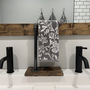 May include: A freestanding towel holder featuring a dark stained wooden base and a vertical black industrial pipe arm. A gray hand towel with a white floral pattern hangs from the pipe. The unit is positioned between two matte black faucets.