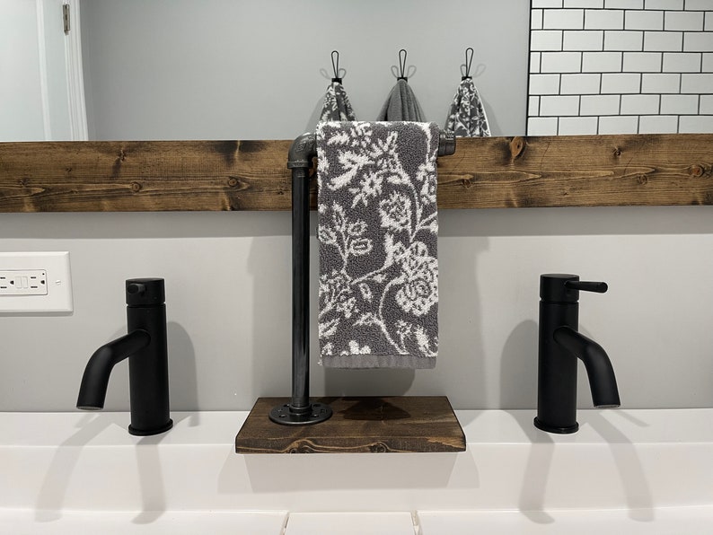 May include: A freestanding towel holder featuring a dark stained wooden base and a vertical black industrial pipe arm. A gray hand towel with a white floral pattern hangs from the pipe. The unit is positioned between two matte black faucets.