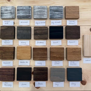 May include: A collection of stained wood samples in various colors, including Antique White, Ebony, and Gray. Each sample is a small, square block with a label underneath indicating the color name. The samples are arranged on a light-colored wooden surface.