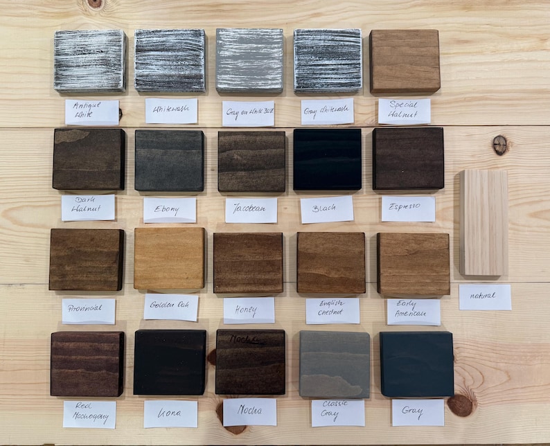 May include: A collection of stained wood samples in various colors, including Antique White, Ebony, and Gray. Each sample is a small, square block with a label underneath indicating the color name. The samples are arranged on a light-colored wooden surface.