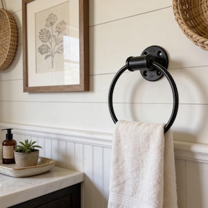 May include: Black metal towel ring with a white towel hanging from it, mounted on a white wall. A framed botanical print and a woven basket are also visible. The bathroom decor has a farmhouse style.