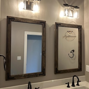May include: Two rectangular mirrors with dark wood frames mounted above a double vanity with white countertops and black faucets. Two wall-mounted light fixtures with clear seeded glass shades are positioned above each mirror. The right mirror features a circular logo with the text Lightrooom.